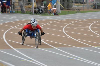 Wheelchair racer in a red shirt and white helmet pushes toward the camera