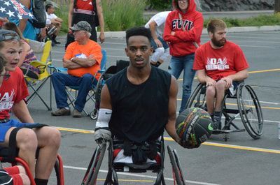 Player in a black tank dribbles beside his wheelchair with spectators behind him