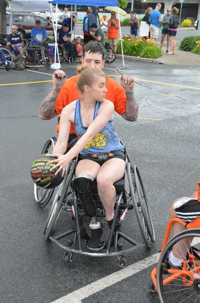 Player in a blue tank holds the ball at her side as a defender in orange guards