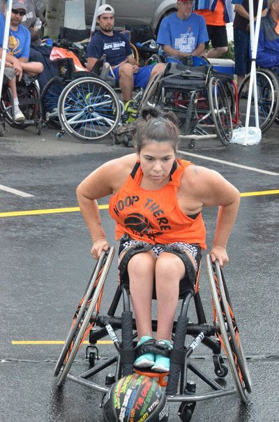 Player in an orange tank leans forward to push her chair, ball in her lap