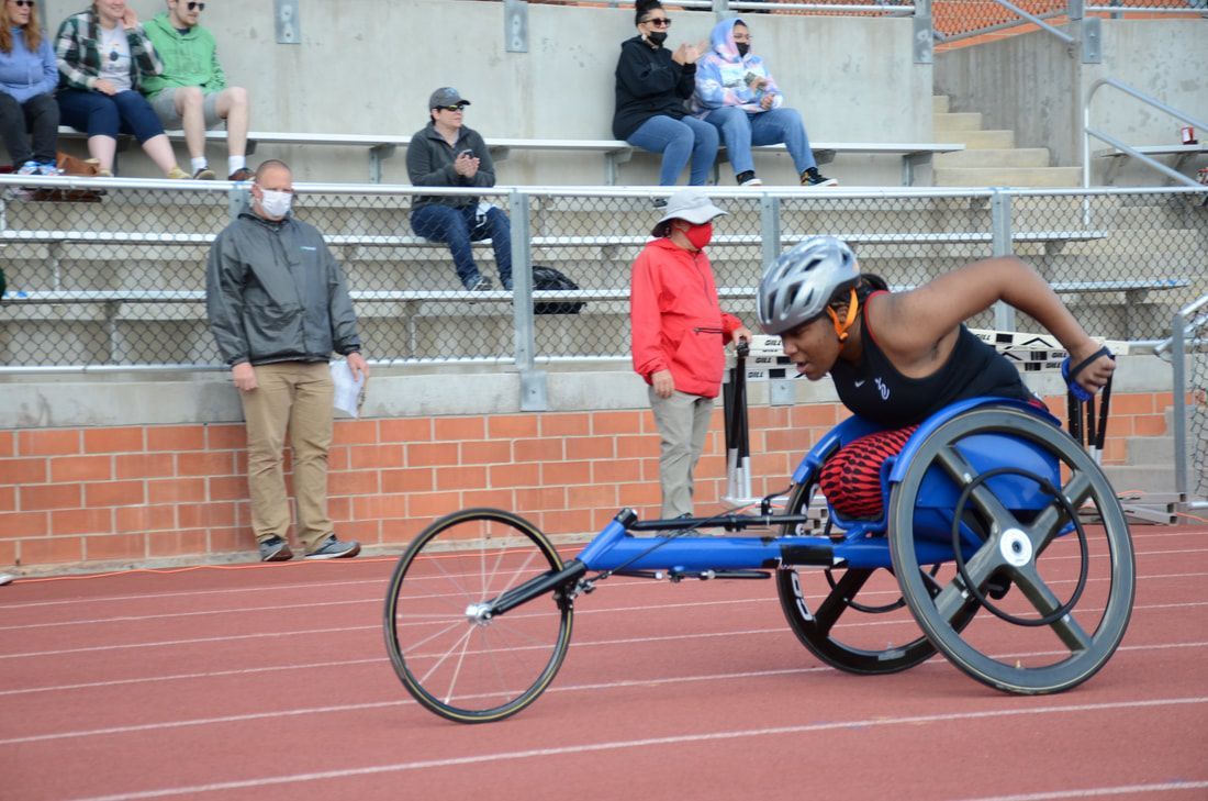 Athlete pushes a blue racing wheelchair past officials and the stands