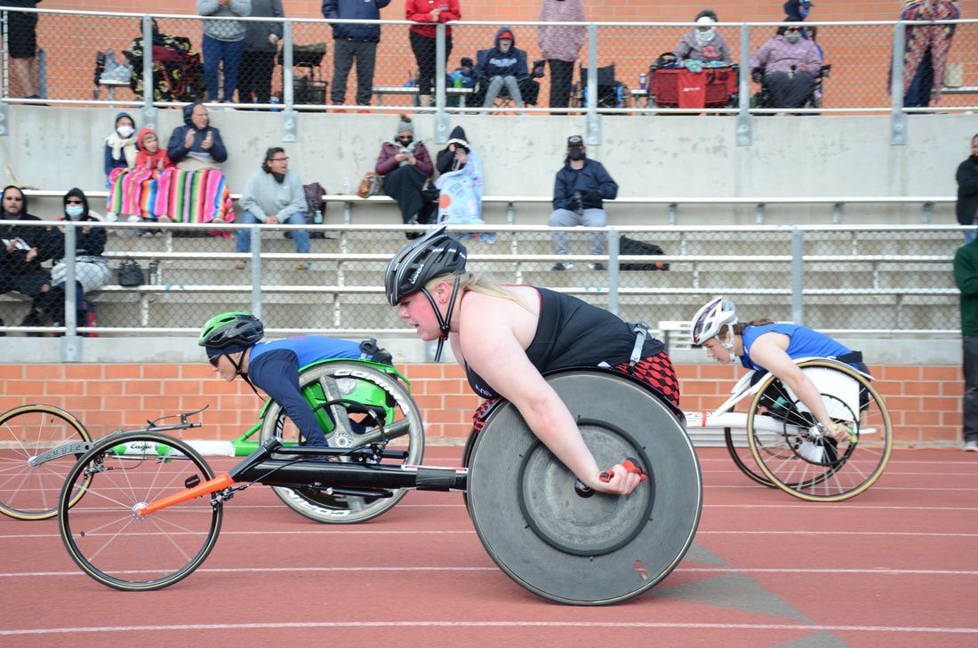 Wheelchair racer with black disc wheels leads two others past the stands