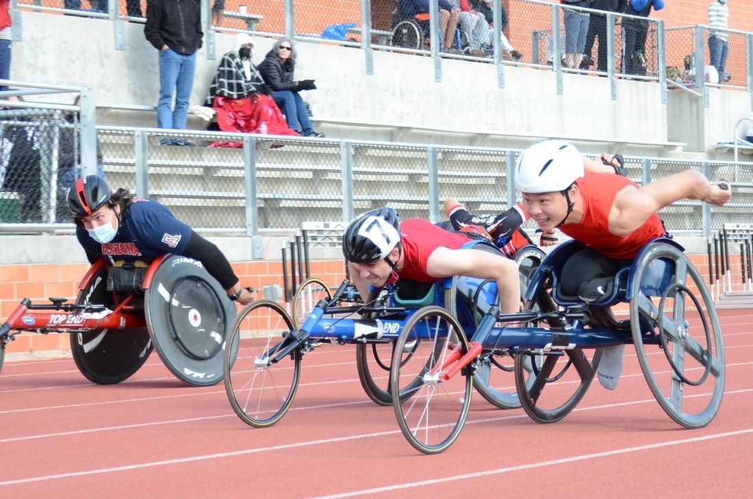 Three wheelchair racers push side by side, one wearing a face mask