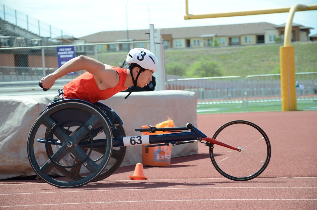 Wheelchair racer in an orange top and white helmet reaches forward in chair 63