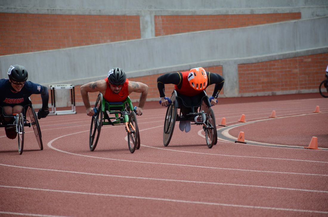 Three wheelchair racers, one in an orange helmet, come off the curve