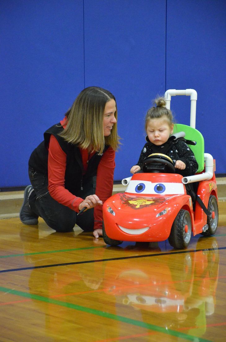 Volunteer kneels beside a toddler driving a red cartoon-face ride-on car
