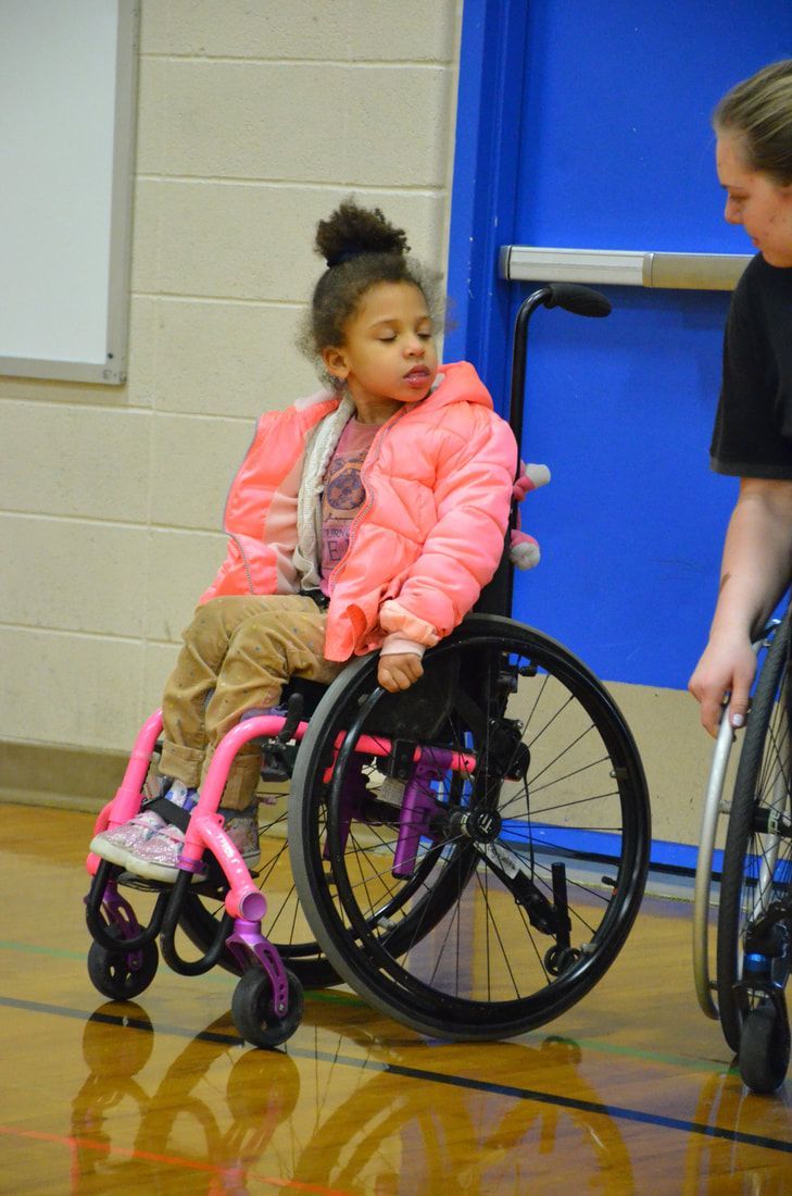 Girl in a pink jacket sits in a pink-framed wheelchair beside a coach