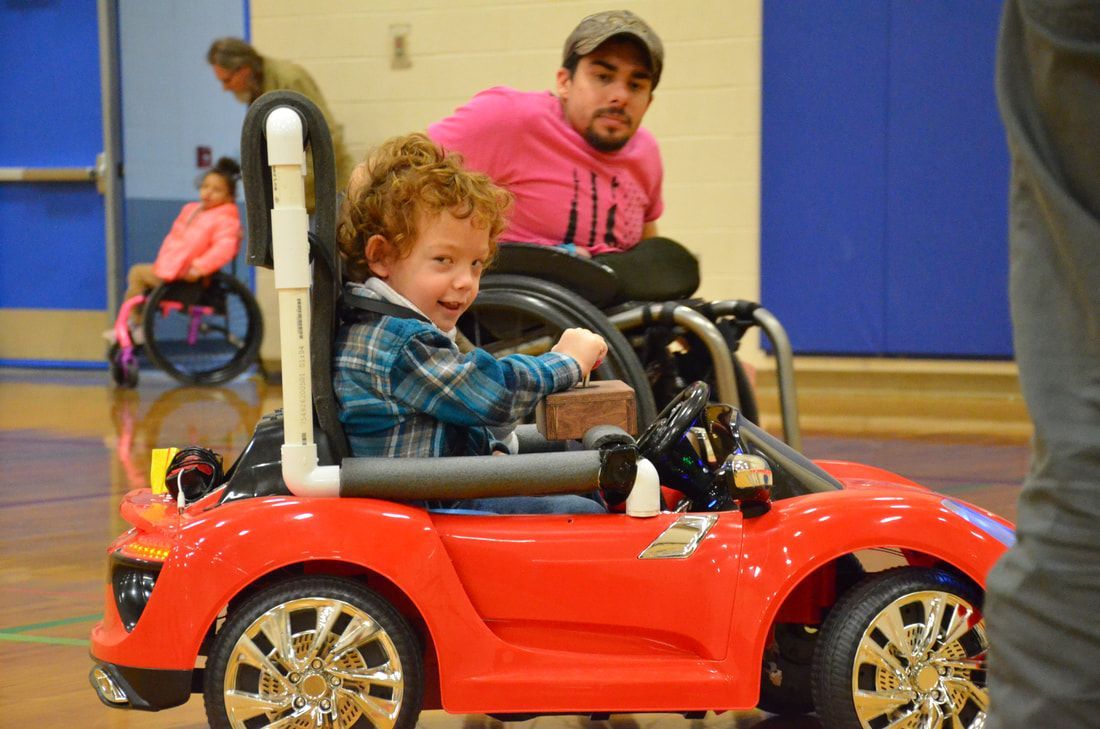 Boy in plaid smiles from a red ride-on car, a wheelchair athlete behind him