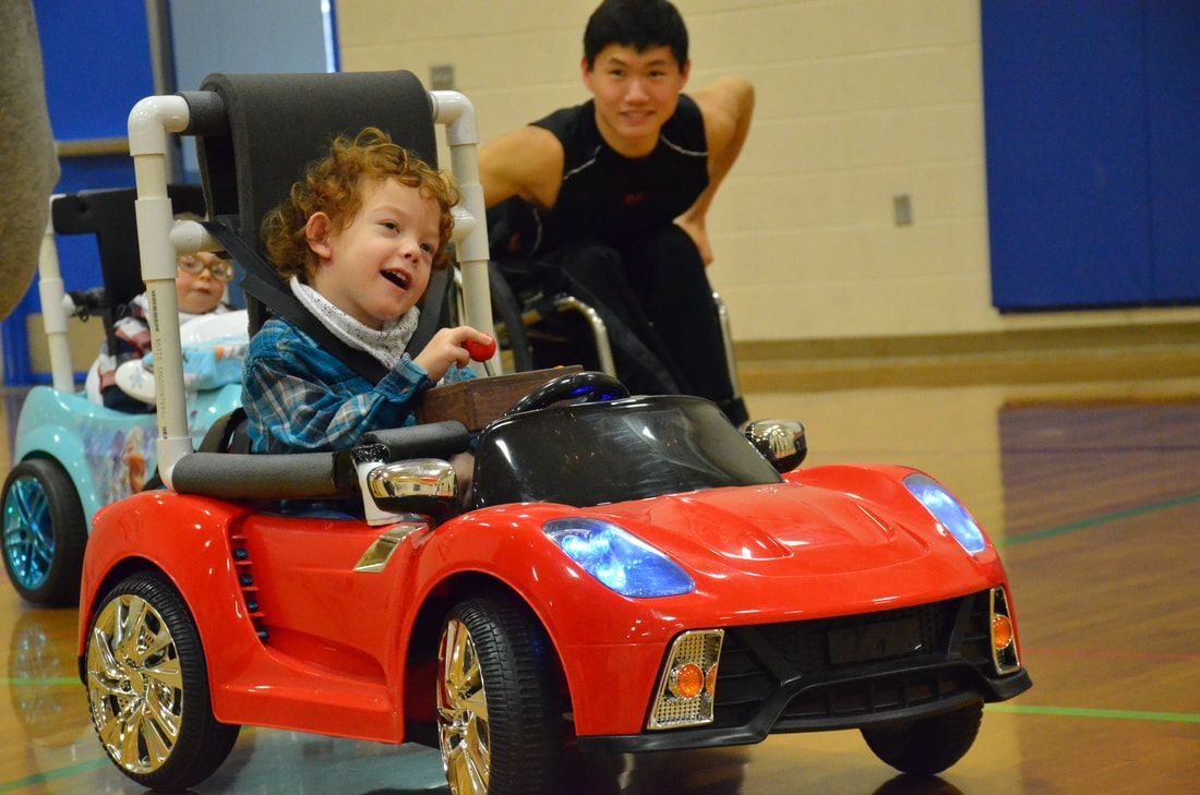 Laughing boy steers a red sports ride-on car as a wheelchair athlete follows