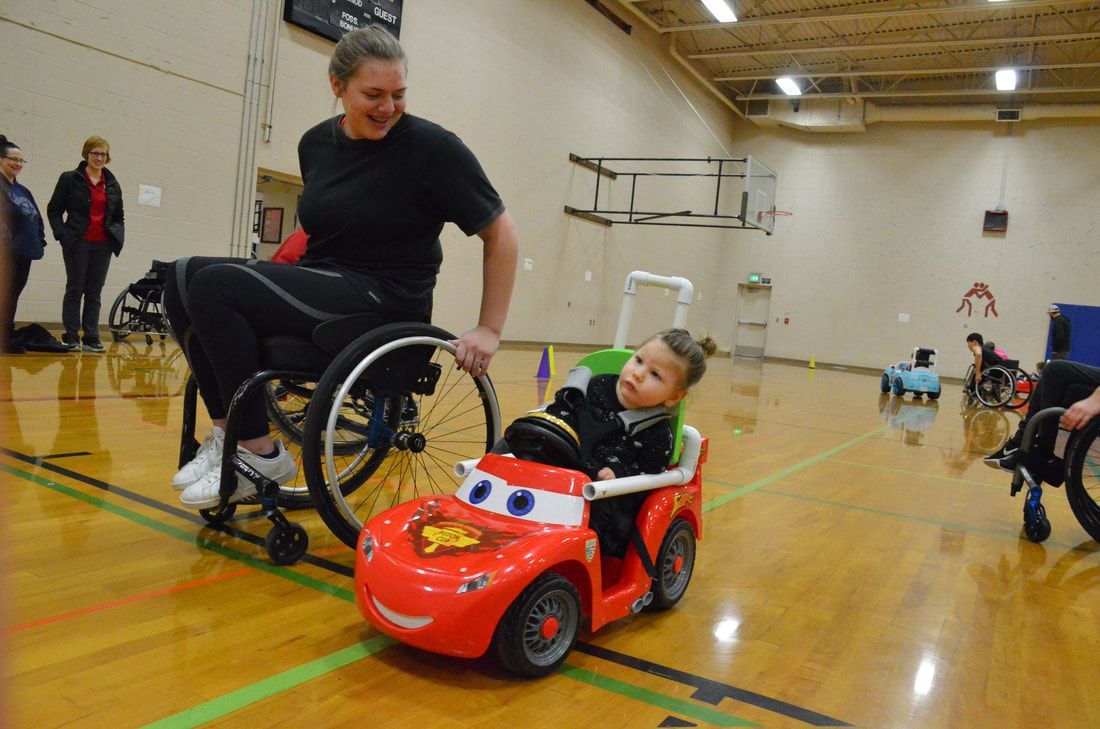 Athlete in a wheelchair rolls beside a toddler in a red cartoon-face car