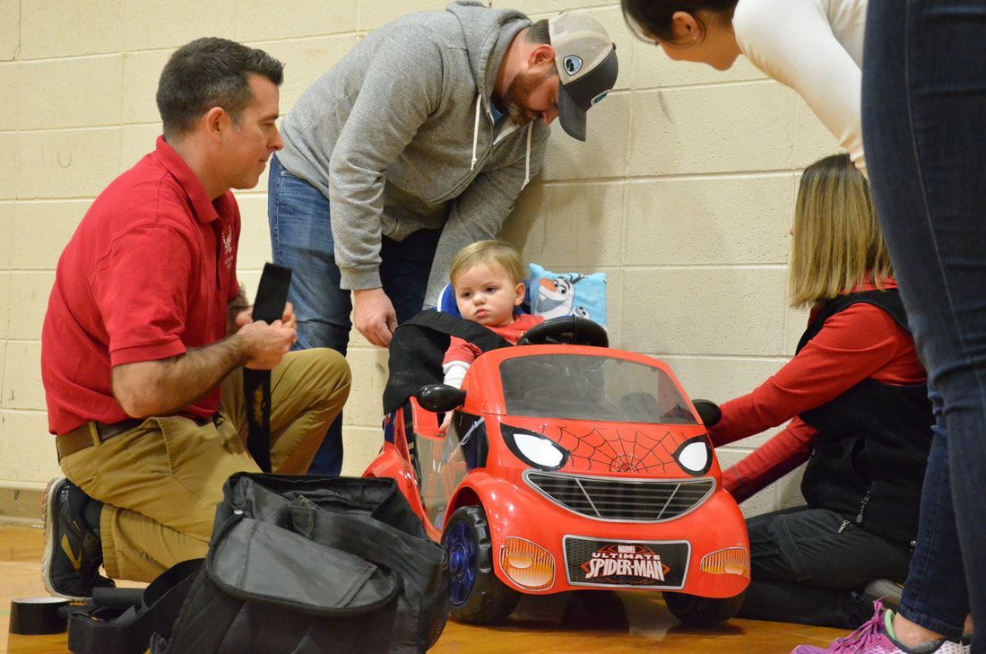 Volunteers fit a toddler into a red Spider-Man ride-on car