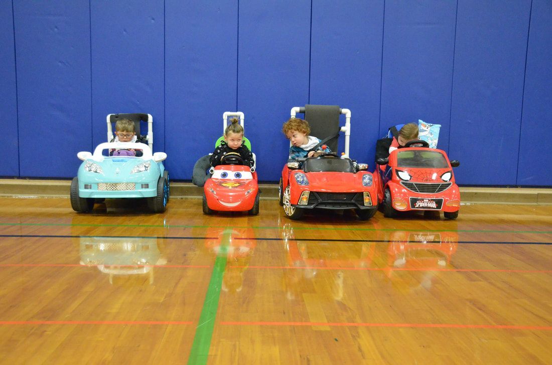 Four children in ride-on cars line up against a blue gym wall