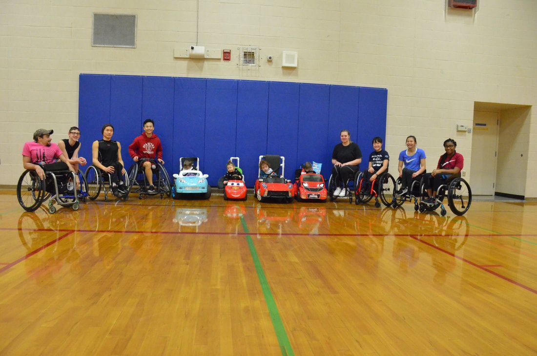 Wheelchair athletes and children in ride-on cars line up across the gym