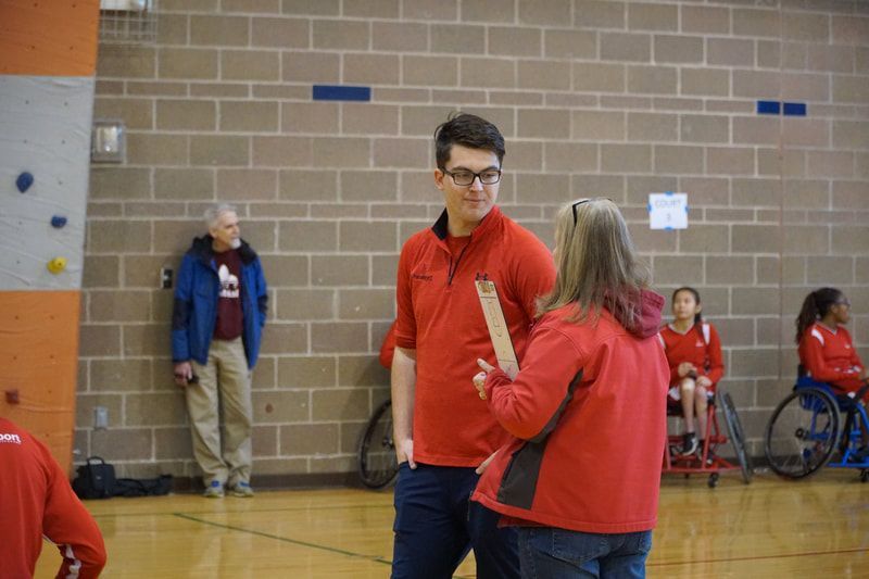 Coach with a clipboard talks with a woman in a red jacket on the sideline