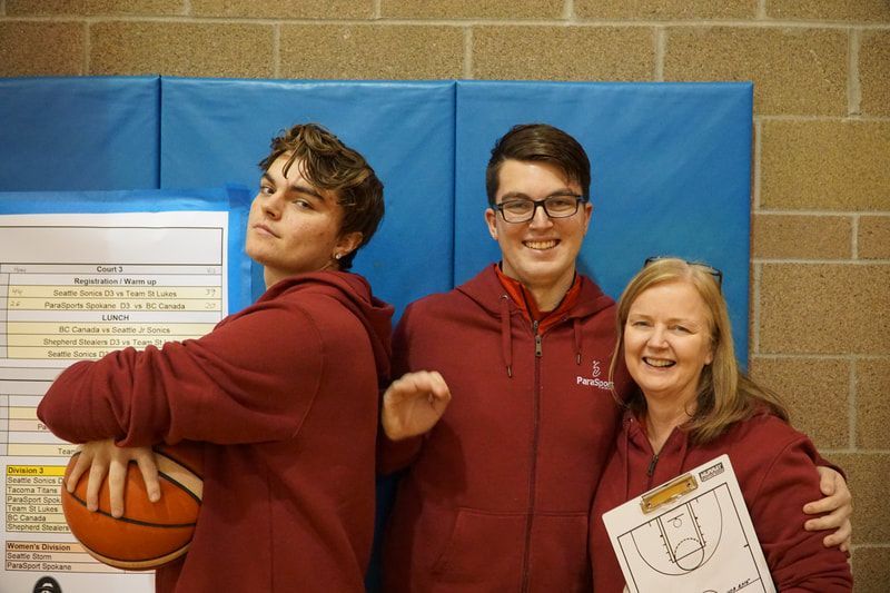 Two players in maroon hoodies pose with a coach holding a clipboard