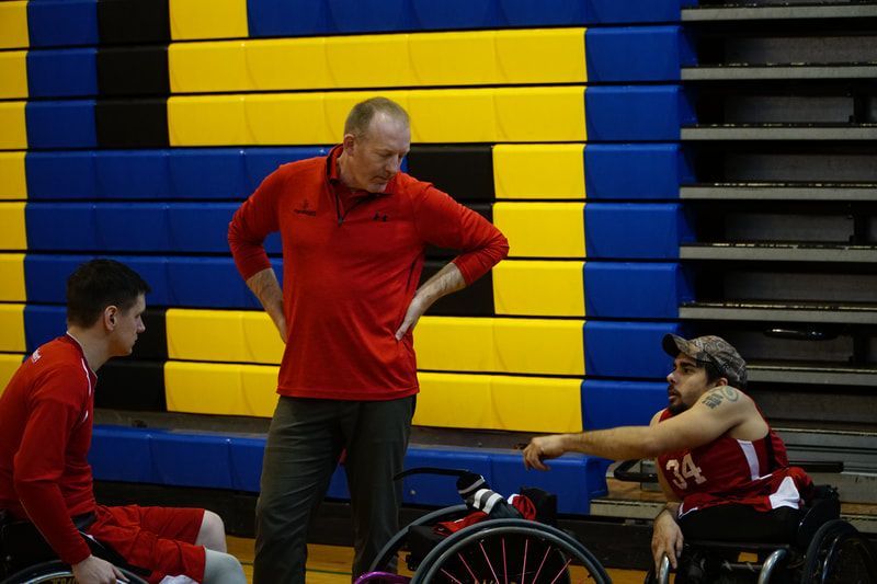 Coach in red with hands on hips talks with players on the bench