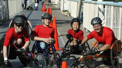 Four racers in red shirts pose in racing wheelchairs on a bridge walkway