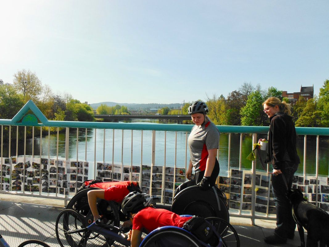 Racers in red shirts and two women gather on a bridge above a river