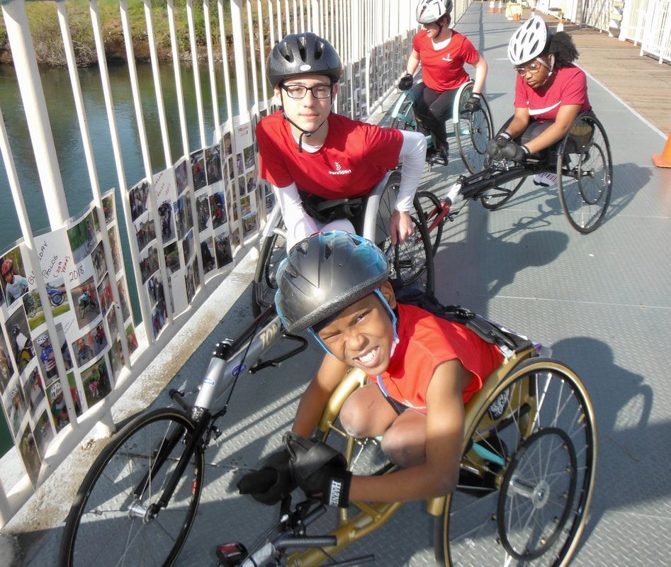 Grinning young racer in a gold chair leads teammates in red across a bridge