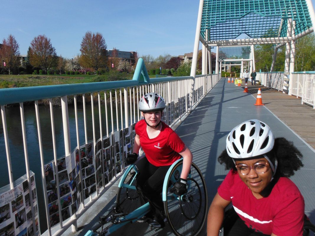 Two smiling athletes in white helmets on a footbridge, one in a teal chair