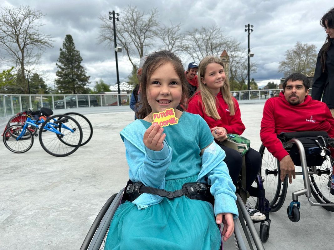 Girl in a teal dress holds a Future Bloomie badge from her wheelchair