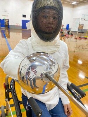Seated parafencer in a mask and white jacket points the blade at the camera