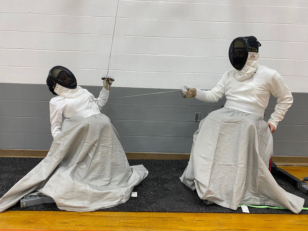 Two parafencers in white gear face off seated on fencing frames