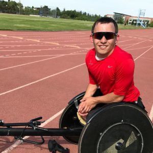 Isaiah Rigo in sunglasses and a red shirt leans on his racing wheelchair
