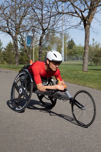 Racer in a white helmet and red shirt pushes a racing chair on a park path