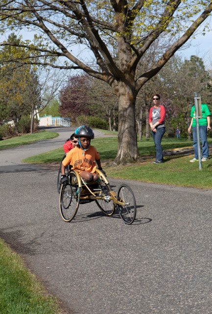 Young athlete in an orange shirt pushes a gold racing chair down a park path