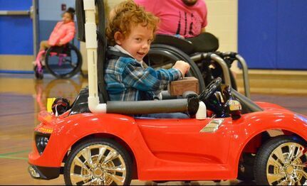 Smiling child drives a red Go Baby Go ride-on car in a gym