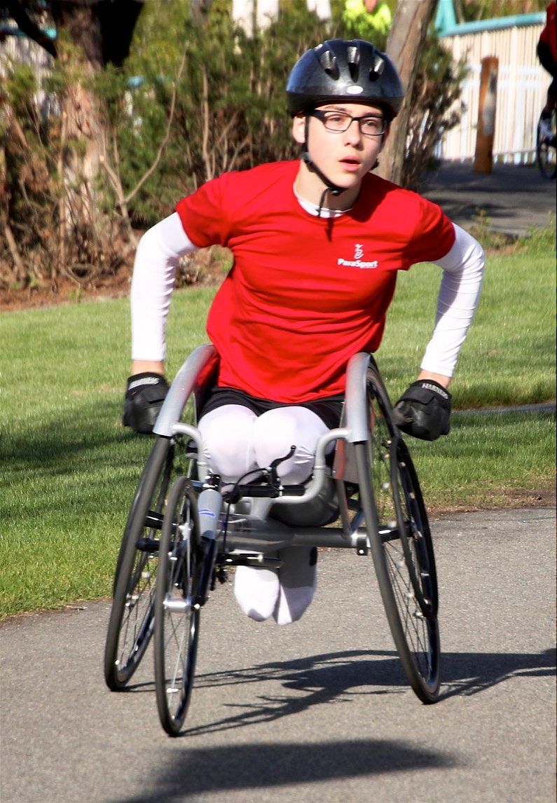 Young racer in a red shirt and glasses pushes a racing chair toward the camera