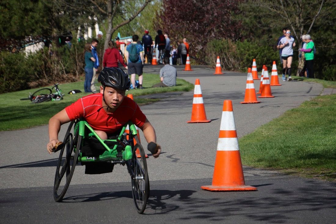 Racer in a red shirt pushes a green racing chair past cones as runners go by