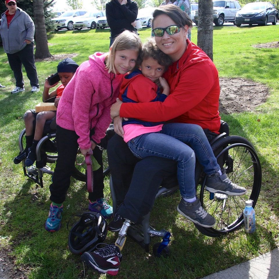 Woman in a red jacket in a wheelchair hugs two children on the grass