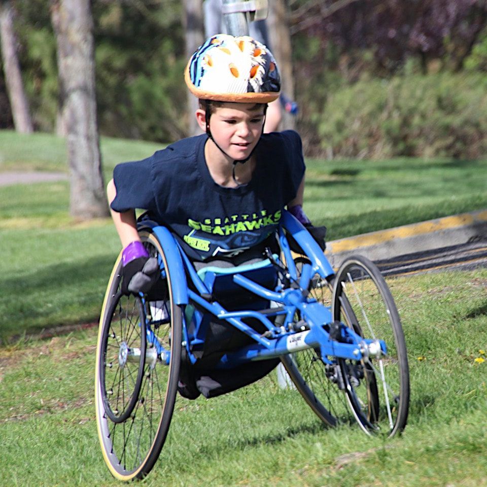 Young racer in a patterned helmet pushes a blue racing chair across the grass