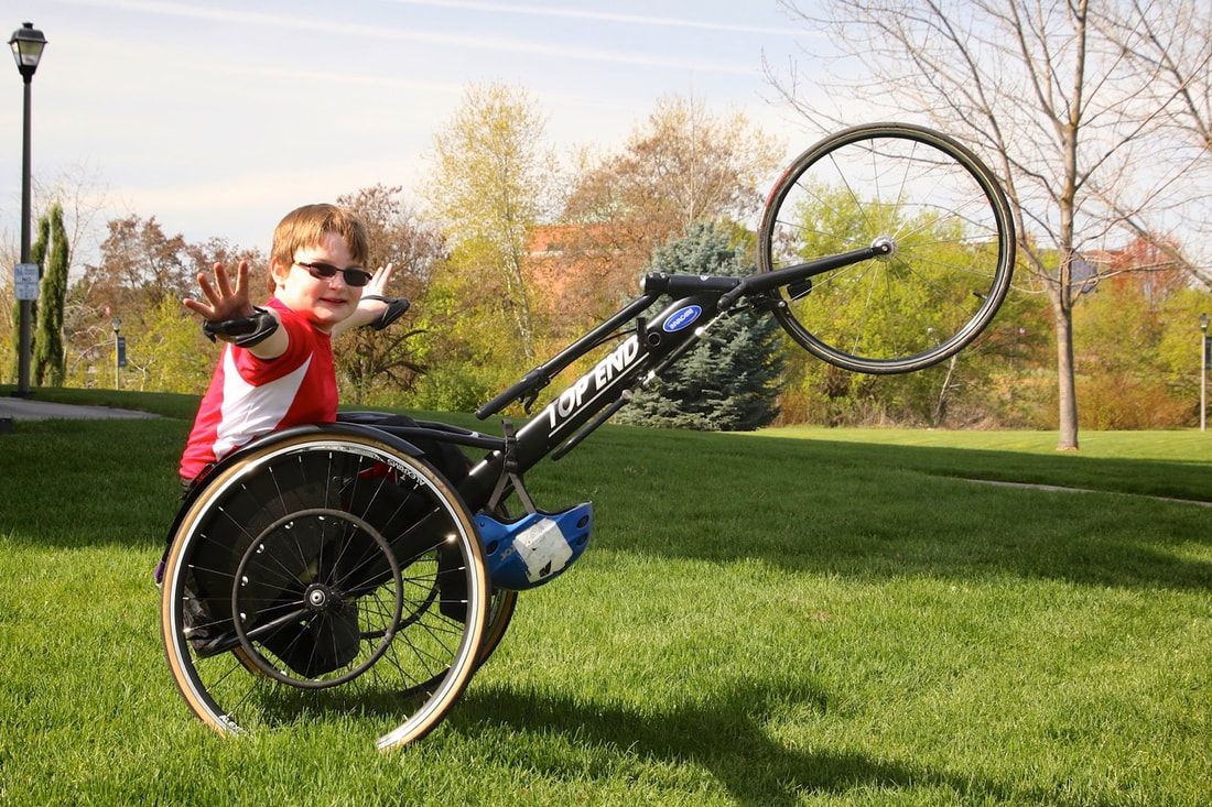 Child in sunglasses waves from a racing chair tipped back, front wheel up