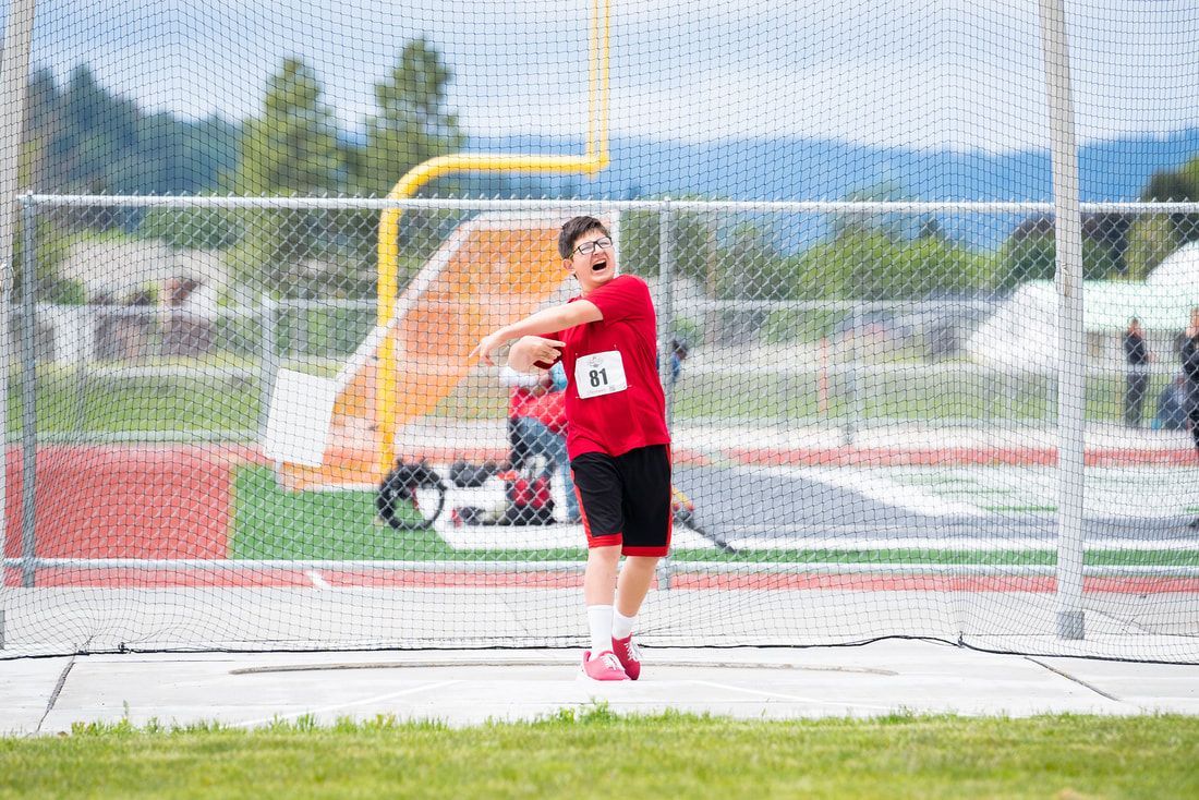 Young athlete in red, bib 81, winds up a throw inside the netted cage