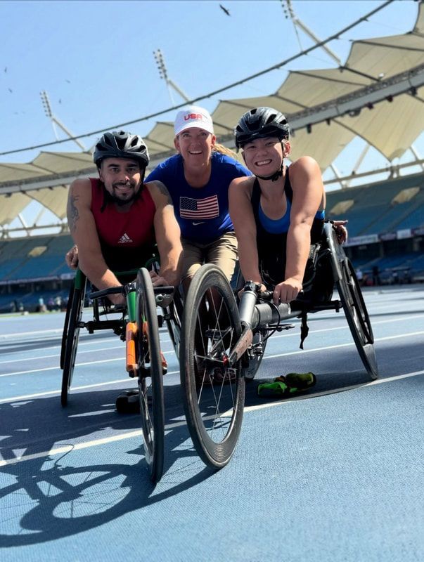 Two wheelchair racers in helmets pose on the track with a woman in a USA cap