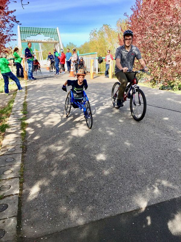 Young racer in a blue chair rides beside a cyclist on a shady trail