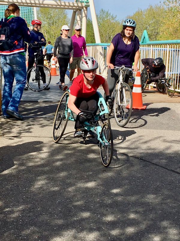 Young racer in a teal racing chair starts out beside a cyclist in purple