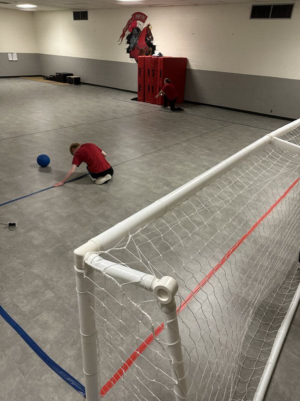 Goalball player in red kneels and reaches across the court near a blue ball