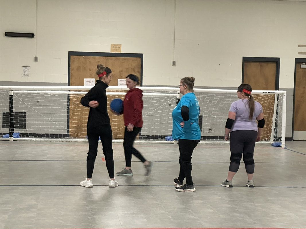 Four goalball players talk in front of the goal net, one holding the ball