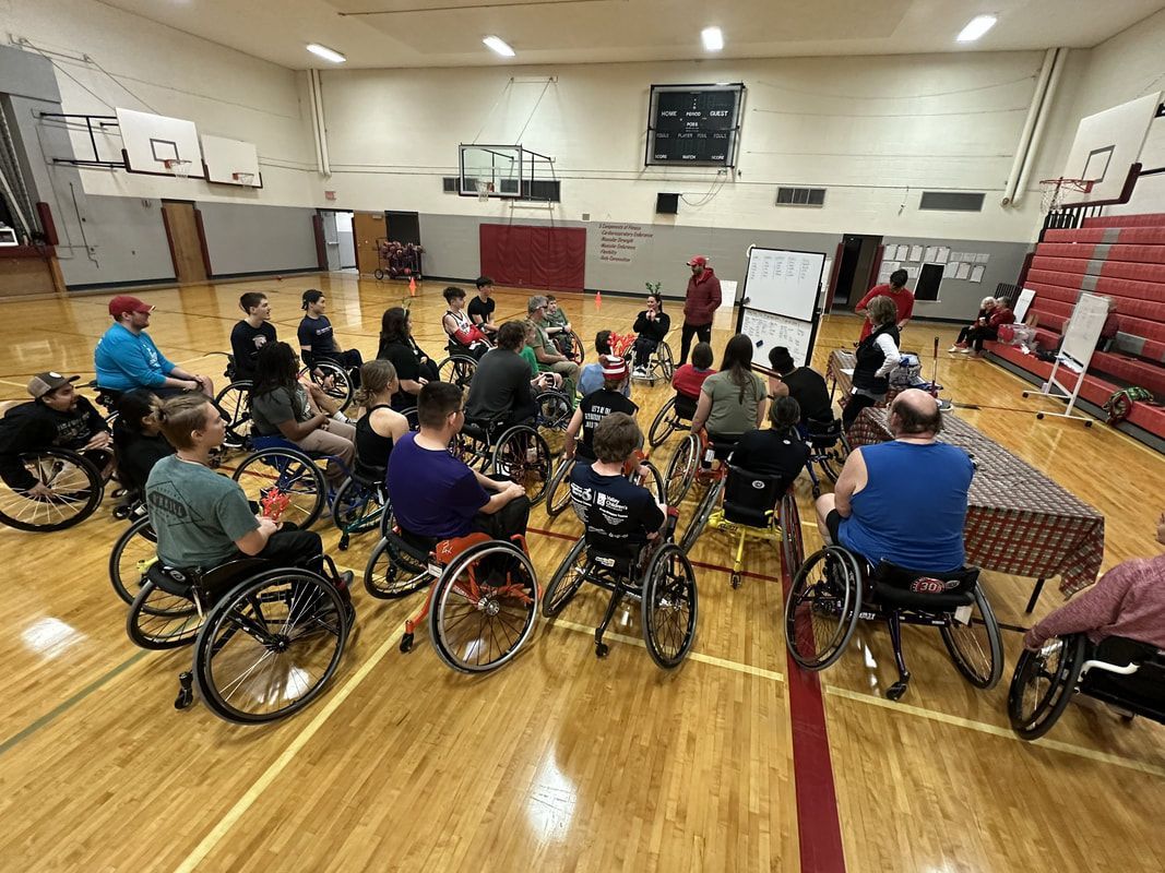 Large group in sport wheelchairs gathers near a whiteboard in the gym