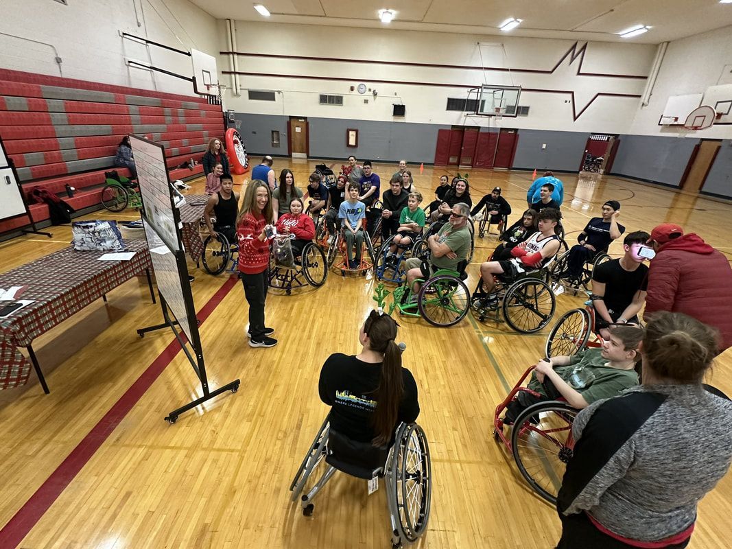 Coach in red talks at a whiteboard to a crowd of athletes in wheelchairs