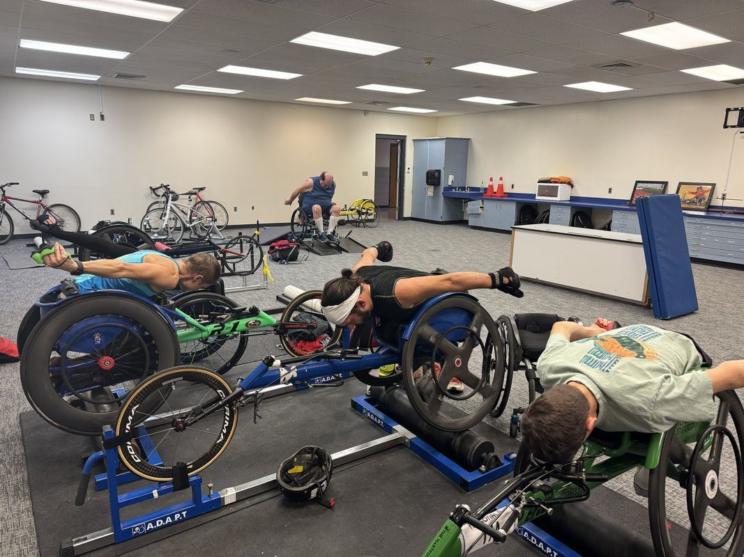 Three athletes train in racing wheelchairs mounted on indoor rollers
