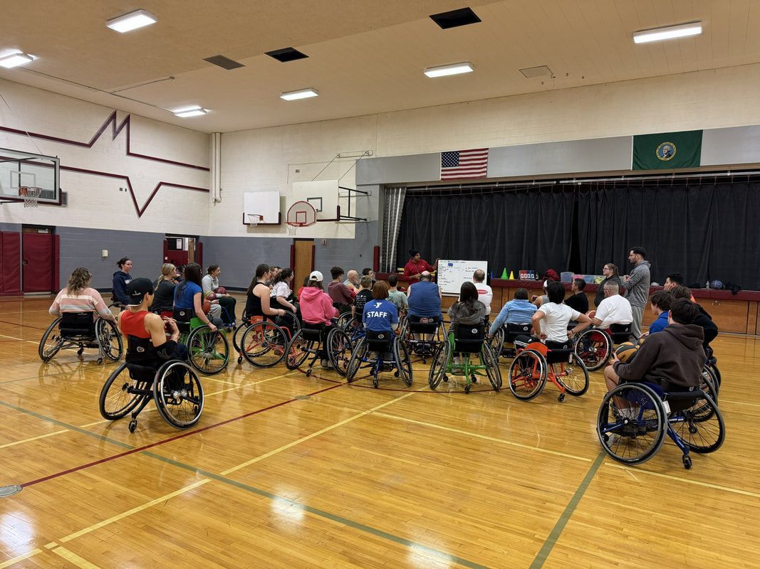 Athletes in sport wheelchairs gather near the stage end of the gym