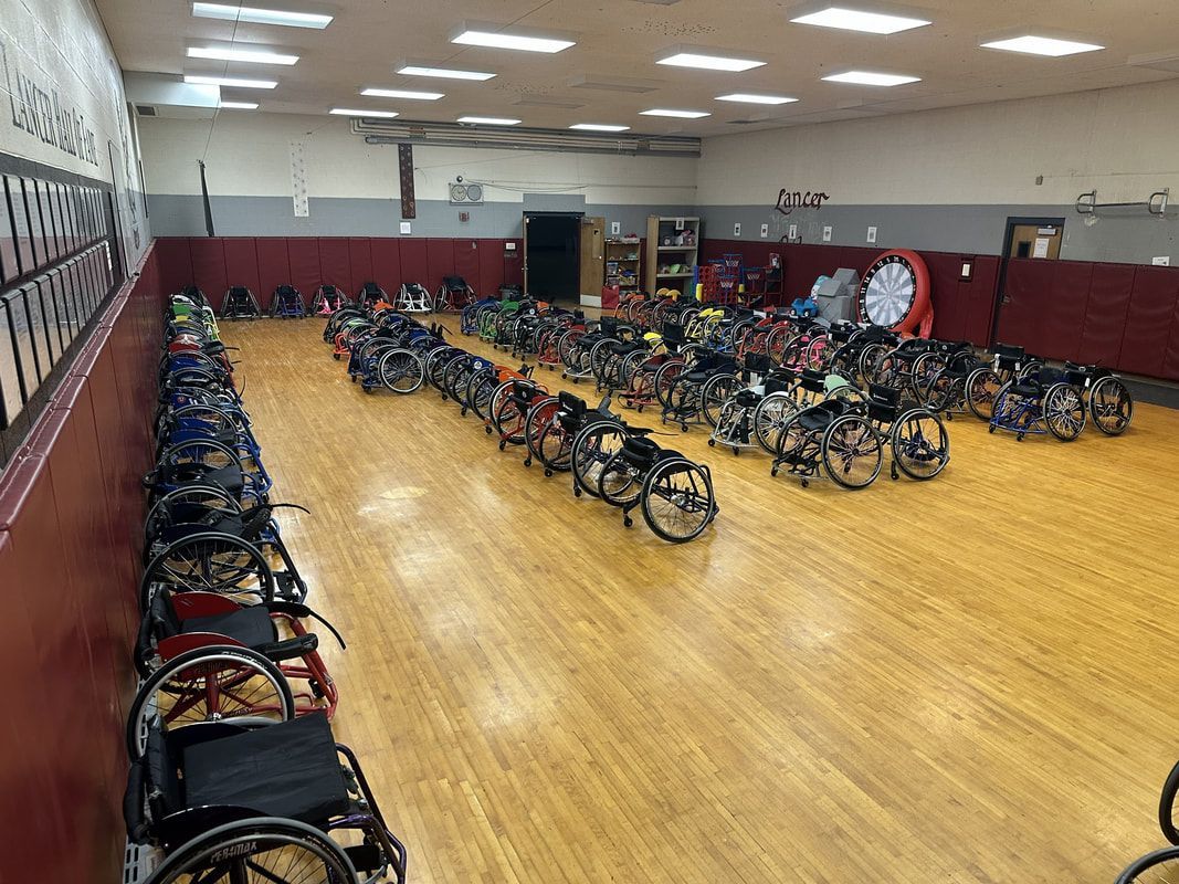 Dozens of sport wheelchairs parked in rows along a long wood-floored room