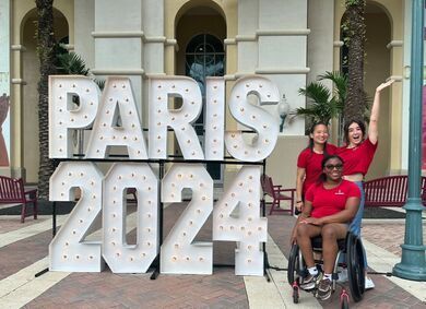 Three athletes in red pose by lighted PARIS 2024 letters, one in a wheelchair