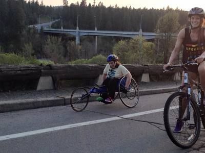Racer in a racing wheelchair and a cyclist on a road with a bridge behind