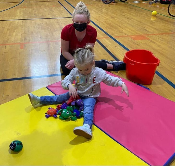 Athlete in red mentors a young child playing with beanbags on a gym mat